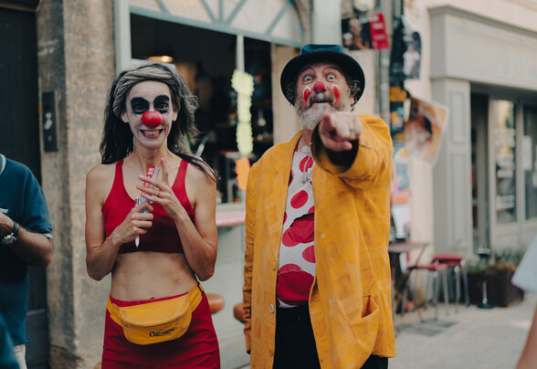 Deux clowns au nez rouge dans une rue, une femme souriante et un homme au chapeau pointant du doigt