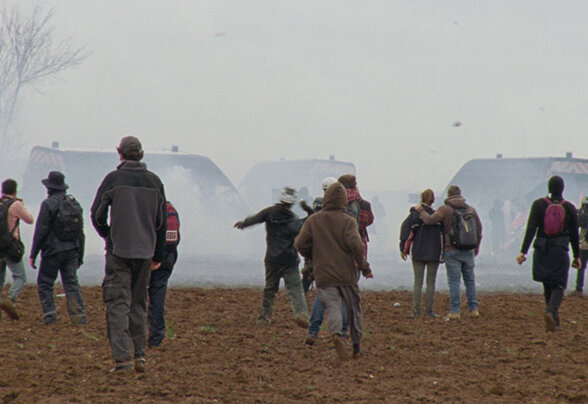 Militants de dos marchant dans un champ labouré vers des fourgons de police, dans un nuage de fumée