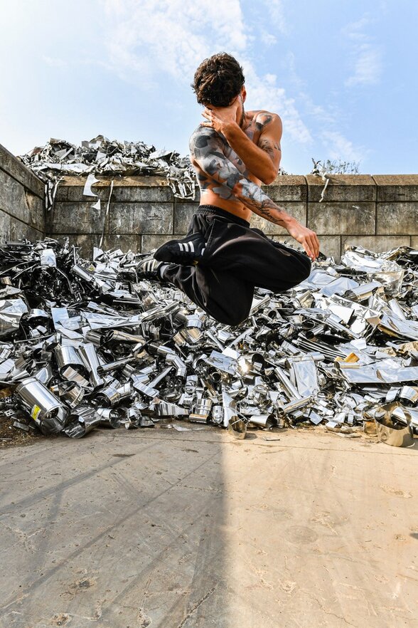 Danseur torse nu au corps peint, en plein saut devant un amas de déchets métalliques brillants